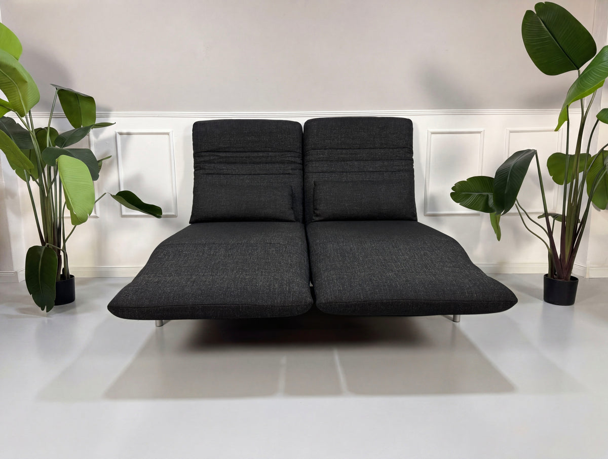 Gebrauchtes graues Rolf Benz Plura Designer Stoff Sofa mit Hocker vor einer Wand, stilvoll und modern eingerichtet.