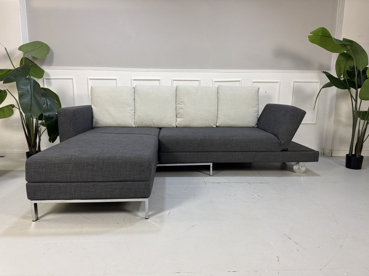Gebrauchtes graues Brühl Fourtwo Designer Stoff Ecksofa vor einer Wand, stilvoll und modern eingerichtet.