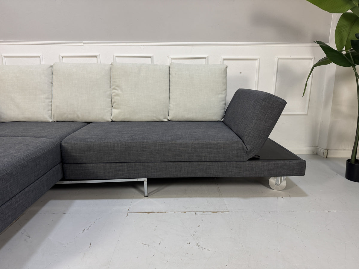 Gebrauchtes graues Brühl Fourtwo Designer Stoff Ecksofa vor einer Wand, stilvoll und modern eingerichtet.
