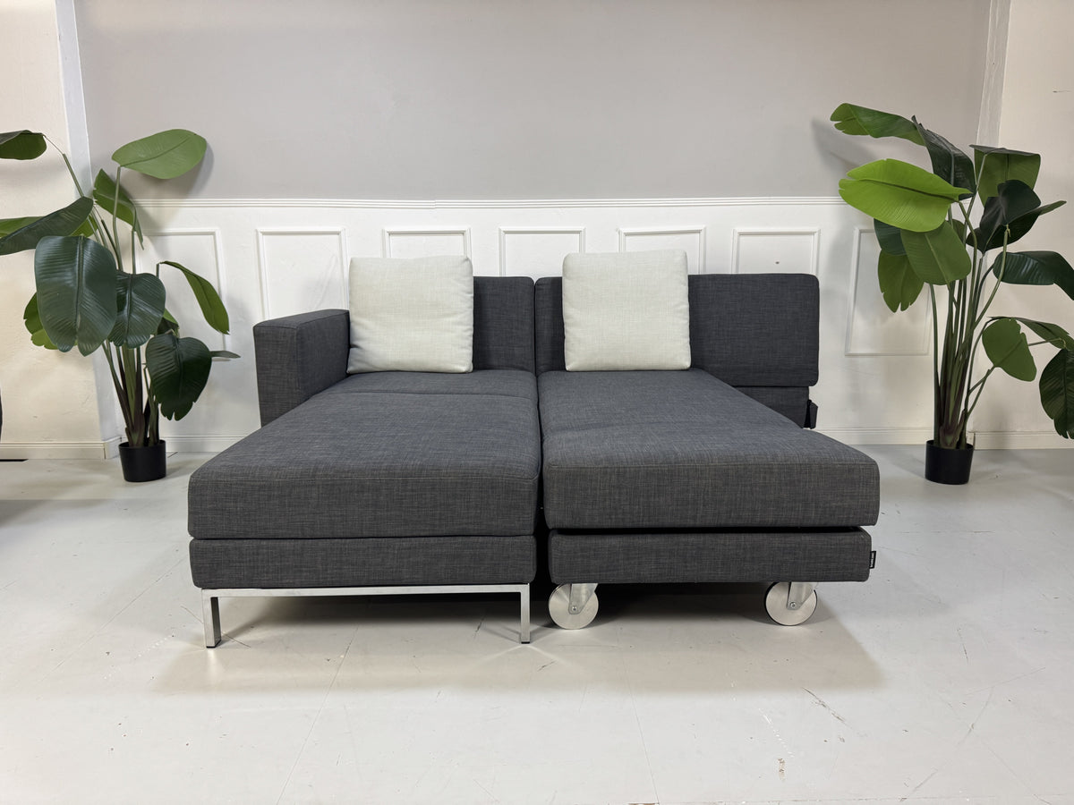 Gebrauchtes graues Brühl Fourtwo Designer Stoff Ecksofa vor einer Wand, stilvoll und modern eingerichtet.